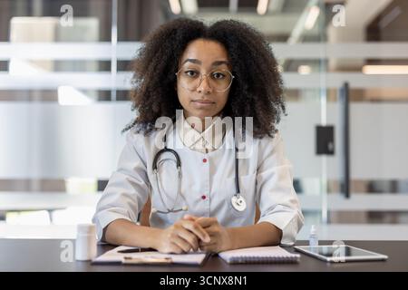 Jeune femme médecin portant des lunettes et stéthoscope assis en toute confiance à table dans le cabinet médical moderne. Médecin apparaît professionnel et attentif, avec des documents et tablette en vue Banque D'Images