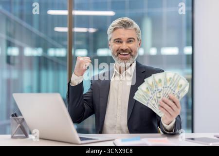 Homme d'affaires mûr en costume serrant un fan de billets de cent dollars et pompant son poing à un bureau couvert d'ordinateur portable. Célébrer la réussite financière. Richesse et réussite entrepreneuriale confiante Banque D'Images