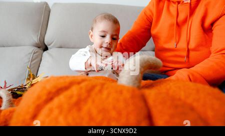 Une mère méconnaissable recadrée est assise avec un petit garçon de 4 mois lors d'une séance photo sur le thème de l'Halloween à la maison. Le bébé est fasciné par une grande citrouille Banque D'Images