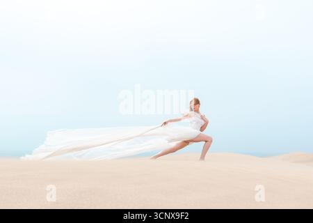 Une jeune femme élégante dans une robe blanche fluide danse gracieusement sur une plage de sable. Le tissu tremble dans la brise, capturant l'essence de l'été Banque D'Images