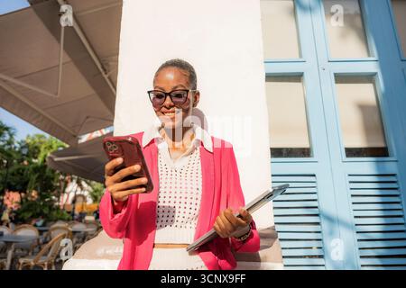 Femme d'affaires noire dans un costume rose souriant tout en utilisant son téléphone et tenant des documents à Cartagena de Indias, Colombie. Un compleme serein avec vue sur la rue Banque D'Images