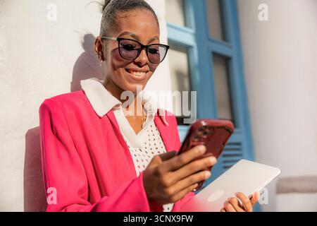 Femme d'affaires noire souriante à Cartagena de Indias, Colombie, dans un blazer rose vif, utilisant son smartphone et sa tablette tout en se tenant dehors, agréable Banque D'Images
