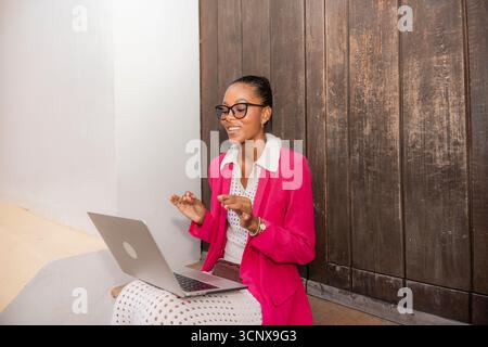 Une femme d'affaires noire professionnelle dans un blazer rose sourit alors qu'elle s'engage dans une session de travail à distance sur son ordinateur portable dans la ville animée de Carthagène de I. Banque D'Images