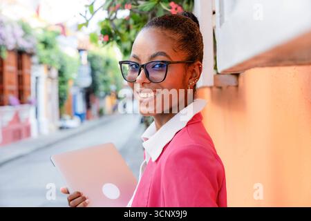 Une femme d'affaires noire confiante dans un blazer rose tient un ordinateur portable tout en souriant dans les rues animées de Cartagena de Indias, Colombie, mettant en vedette le professeur Banque D'Images