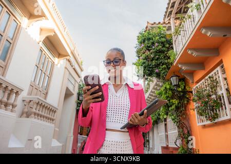 Femme d'affaires noire confiante dans un costume rose vif utilise son smartphone et tient une tablette tout en souriant dans une charmante rue de Cartagena de Indias, Banque D'Images