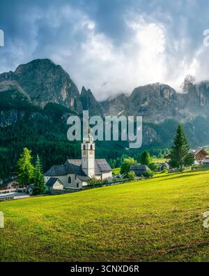 Église alpine dans le village de Colfosco avec des sommets Dolomites spectaculaires en toile de fond, des prairies vertes et un ciel nuageux. Paysage de montagne du Tyrol du Sud, Italie Banque D'Images