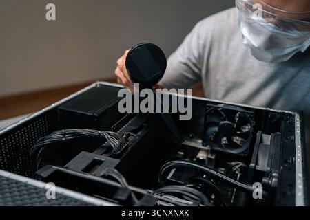 Technicien en informatique portant un masque facial et des lunettes de protection à l'aide d'un aspirateur pour enlever la poussière et les débris de l'intérieur du boîtier de l'ordinateur Banque D'Images
