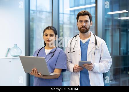 Médecin masculin dans la blouse de laboratoire et infirmière féminine dans les gommages se tiennent dans la clinique tenant la tablette et l'ordinateur portable, mettant en valeur le travail d'équipe médical moderne, les outils numériques et les progrès technologiques dans les soins de santé Banque D'Images