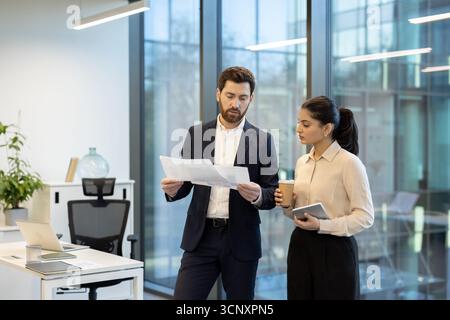 Deux hommes d'affaires divers discutent de documents importants et de données financières, collaborant à un projet dans un environnement de bureau contemporain lumineux Banque D'Images