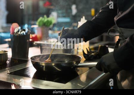 Homme caucasien d'âge moyen portant des gants préparant la nourriture dans la cuisine professionnelle, tenant des ustensiles de cuisine et remuant les ingrédients dans une poêle à frire sur la cuisinière, vapeur montant de la poêle Banque D'Images