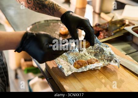 Jeune femme adulte caucasienne au bras tatoué portant des gants noirs préparant des pommes de terre cuites au four en papier d'aluminium sur planche de bois dans la cuisine professionnelle, les mains organisant soigneusement la nourriture pour servir Banque D'Images