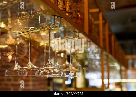 Verres à vin vides suspendus à l'envers du support dans le bar du restaurant, reflétant la lumière ambiante chaude, verrerie disposée en rangée nette, arrière-plan flou avec mur de briques visible Banque D'Images