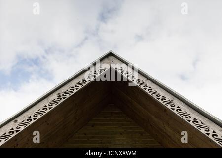Vue rapprochée d'un pignon de toit en bois avec des garnitures décoratives complexes, pointant vers le ciel. Le bois chaud contraste avec un ciel bleu nuageux, transportant Banque D'Images