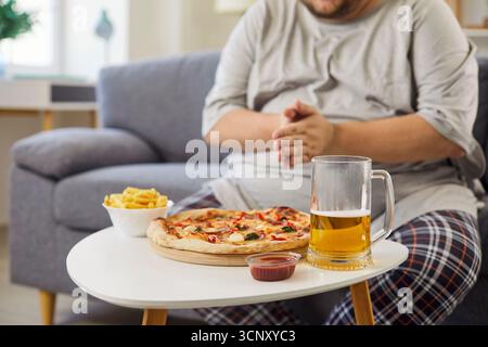 Homme en surpoids sur le canapé avec pizza, frites et bière, rompre le régime alimentaire à la maison Banque D'Images