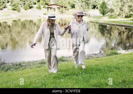 Deux amies seniors marchant ensemble à l'extérieur dans la nature près du lac, tenant les mains sur l'herbe verte, des tenues élégantes avec des chapeaux, âge positif lifestyl Banque D'Images