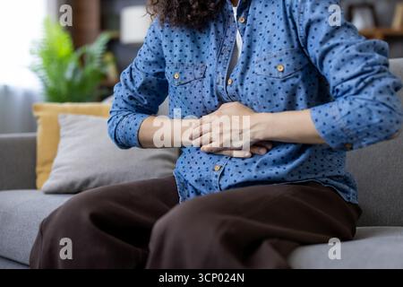 Femme assise sur un canapé à la maison aux prises avec un malaise aigu à l'estomac et des douleurs au ventre, serrant son abdomen en raison de l'indigestion, des menstruations ou d'autres problèmes de santé Banque D'Images