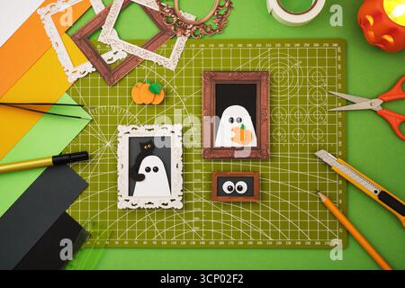 Artisanat en papier d'Halloween avec des découpes fantômes et citrouilles dans des cadres décoratifs, entourés de papier coloré, d'outils, de ciseaux, de cutter et de bougie sur un gre Banque D'Images