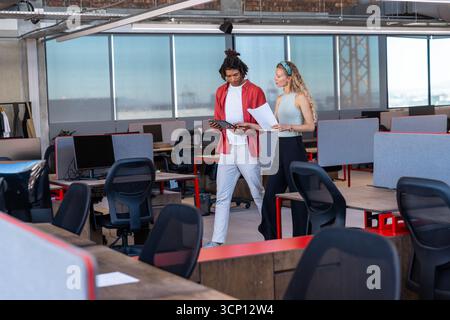 Divers collègues marchent tout en consultant des tablettes et des documents imprimés dans un bureau ouvert Banque D'Images