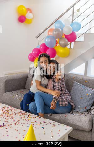 Maman diversifiée et fille adolescente embrassant sur un canapé gris dans le salon avec des confettis, des ballons Banque D'Images
