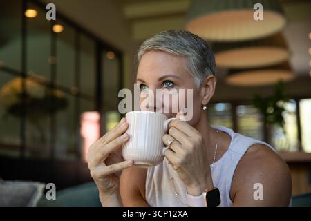 Femme d'âge moyen sirotant de tasse côtelée alors que dans smartwatch et collier à la table de café Banque D'Images