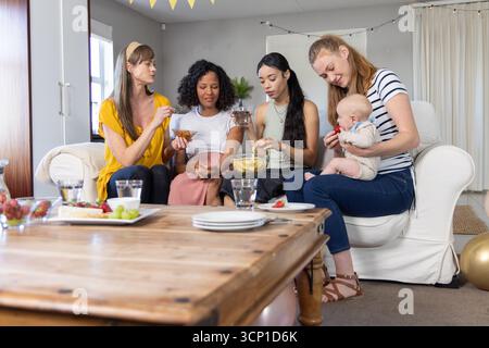 Diverses amies féminines assises dans le salon tenant bébé, mangeant des collations autour de la table basse Banque D'Images