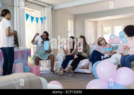 Diverses amies féminines assises sur le canapé ouvrant la combinaison avec des ballons, des bannières dans le salon Banque D'Images