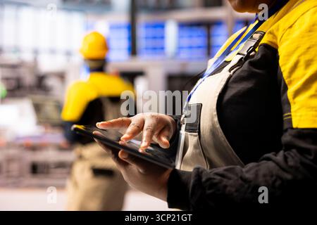 Gros plan des mains interagissant avec l'écran de la tablette dans l'usine intelligente, représentant la transformation numérique dans la fabrication. Le travailleur flou dans l'équipement de sécurité est vu en arrière-plan de l'espace de travail de haute technologie. Banque D'Images