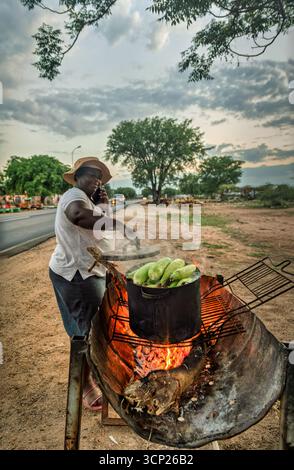Vendeur de rue africain, femme vendant du maïs bouilli sur le feu dans une casserole sur le grill sur le bord de la route dans le canton du village Banque D'Images