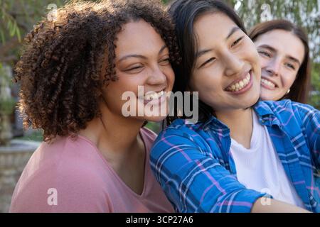 Diverses amies féminines souriant et prenant selfie dans le jardin avec fontaine en pierre et smartphone Banque D'Images