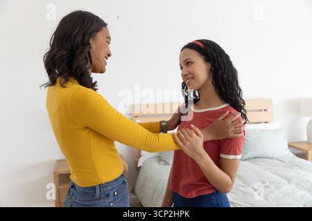 Maman diversifiée et fille adolescente debout à côté d'un lit en bois dans la chambre, plaçant les mains sur les épaules Banque D'Images