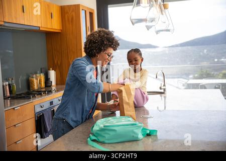 Mère diversifiée avec fille afro-américaine emballant le déjeuner à l'île de cuisine avec sac en papier brun Banque D'Images