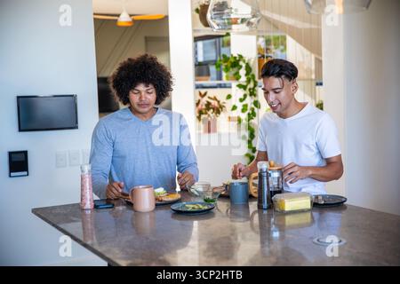 Divers amis masculins préparant et mangeant le petit déjeuner à l'îlot de cuisine en pierre avec des tasses à pain d'avocat Banque D'Images