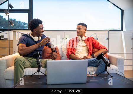 Divers co-hôtes masculins portant des écouteurs et parlant dans des microphones sur le canapé dans la salle d'enregistrement Banque D'Images