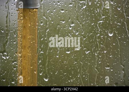 Gros plan de gouttes de pluie sur une vitre, à côté d'un poteau jaune, créant une atmosphère sereine et tranquille. Banque D'Images