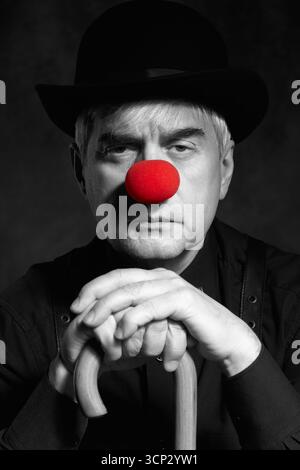 Portrait couleur sélective dramatique d'un clown appuyé sur la poignée d'un parapluie. Un homme âgé avec un nez de clown rouge et un chapeau melon sur la tête. Habillé Banque D'Images