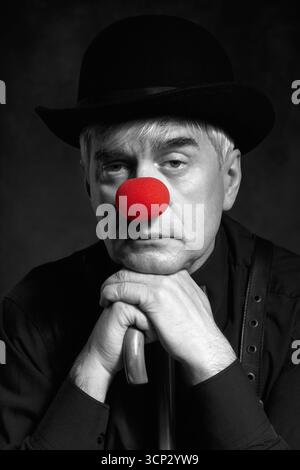 Portrait couleur sélective dramatique d'un clown appuyé sur la poignée d'un parapluie. Un homme âgé avec un nez de clown rouge et un chapeau melon sur la tête. Habillé Banque D'Images