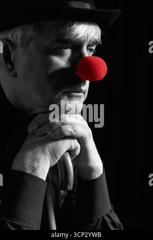 Portrait couleur sélective dramatique d'un clown appuyé sur la poignée d'un parapluie. Un homme âgé avec un nez de clown rouge et un chapeau melon sur la tête. Habillé Banque D'Images