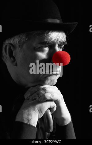 Portrait couleur sélective dramatique d'un clown appuyé sur la poignée d'un parapluie. Un homme âgé avec un nez de clown rouge et un chapeau melon sur la tête. Habillé Banque D'Images