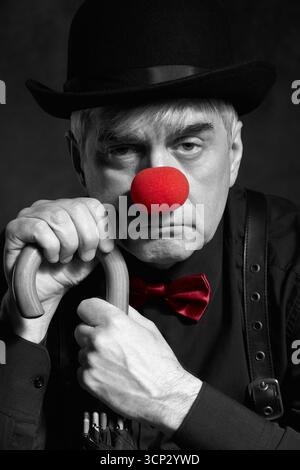Portrait couleur sélective dramatique d'un clown appuyé sur la poignée d'un parapluie. Un homme âgé avec un nez de clown rouge et un chapeau melon sur la tête. Habillé Banque D'Images