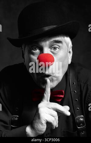 Portrait couleur sélective d'un clown avec le doigt sur les lèvres sur un fond sombre. Un homme âgé avec un nez de clown rouge et un chapeau melon sur la tête. Habillé Banque D'Images