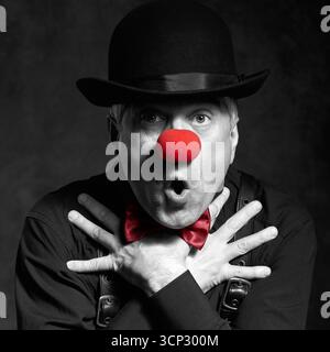 Portrait couleur sélective dramatique d'un clown surpris sur un fond sombre avec les mains sur la poitrine. Un homme âgé avec le nez de clown rouge et bowler ha Banque D'Images