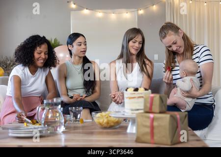 Diverses amies féminines nourrissant des fruits de bébé, discutant sur le canapé à la maison autour du gâteau de célébration Banque D'Images