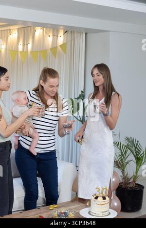 Diverses amies féminines avec bébé célébrant 30e anniversaire autour de la table basse à la maison, gâteau Banque D'Images