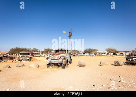 Cimetière de voitures de Solitaire en Namibie - 27 août 2025 : caractéristiques de voitures anciennes rouillées près de petits bâtiments du désert, contre des dunes et des montagnes, CA Banque D'Images