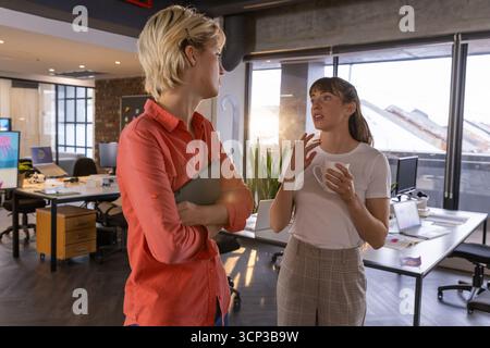 Collègues féminins blancs debout et conversant dans le bureau tenant un ordinateur portable et une tasse à café Banque D'Images