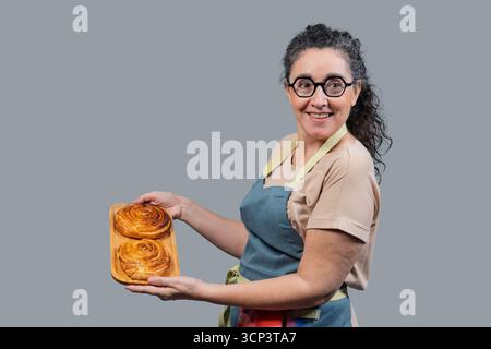Femme dans un tablier présentant une pâte à la cannelle fraîchement cuite sur un plateau en bois Banque D'Images