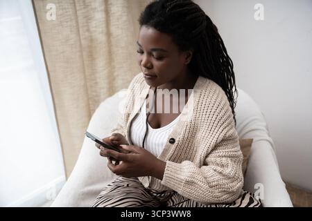Une femme afro-américaine se détend dans son appartement confortable, concentrée sur son smartphone. Banque D'Images