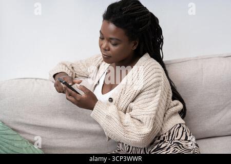 Une jeune femme noire aux cheveux tressés se détend sur son canapé, en s’engageant avec son smartphone. Banque D'Images