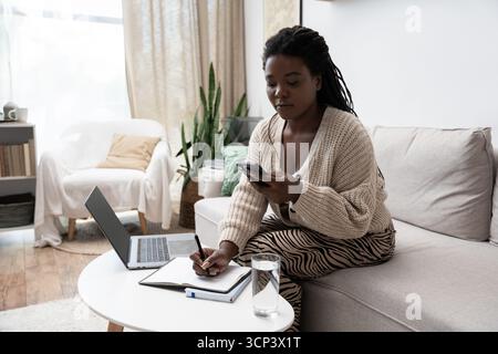 Une femme aux cheveux tressés se détend dans son salon confortable, notant des choses sur son téléphone. Banque D'Images
