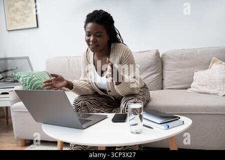 Une femme aux cheveux tressés est assise confortablement, parlant avec enthousiasme dans sa maison moderne. Banque D'Images